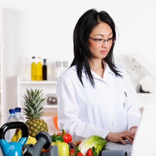 Female dietician in a white coat stands next to a table with a variety of fresh fruit and vegetables including a pineapple